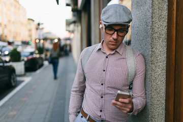 Fashionable retro dressed man with cap, suspenders and eyeglasses standing on city street and using smart phone.