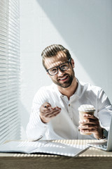 smiling businessman in eyeglasses sitting at workplace with laptop and paperwork on coffee break