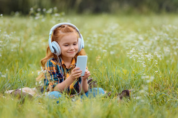 smiling child in headphones using smartphone while resting on blanket in meadow with wild flowers