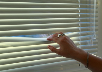 The woman's hand bends the rail of the horizontal blinds to look out the window.
