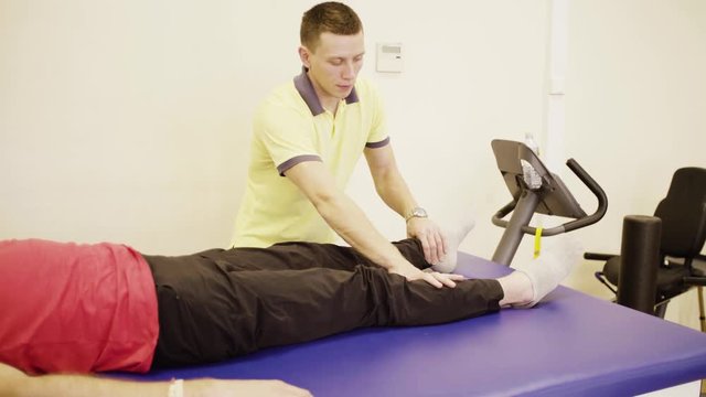 Doctor physiotherapist doing leg exercises for young disabled man at the rehabilitation center. Health reductive gymnastics.