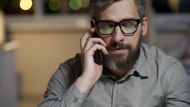 Medium shot of tired mature man in glasses having phone conversation with colleague when sitting at office desk and working late