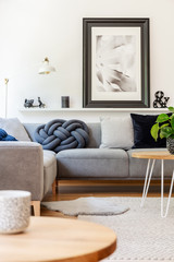 Real photo of handmade knot pillow placed on grey corner sofa standing in white living room interior with lamp, decor and poster on shelf