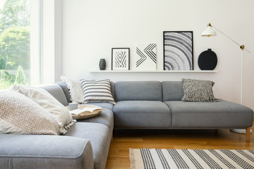 Real photo of white Scandi sitting room interior with metal lamp, corner sofa with cushions and modern art posters