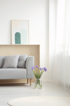 Blue flowers next to grey couch with cushions in simple living room interior with poster. Real photo