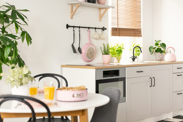 Real photo with blurred foreground of white kitchen interior with pink utensils, window with blinds...
