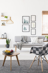 Armchair and wooden end table with fruits standing on carpet in real photo of white living room interior with posters on wall and grey lounge
