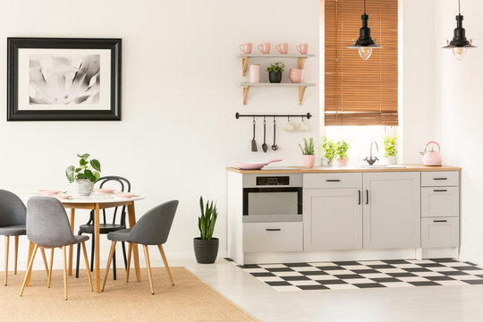 Real photo of open space kitchen interior with dining table with chairs, window with wooden blinds and pink accessories on countertop