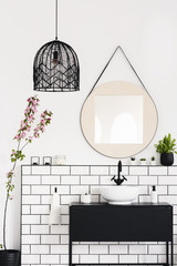 Real photo of a mirror on the wall, black lamp, wash basin with a cupboard and flower in a bathroom interior