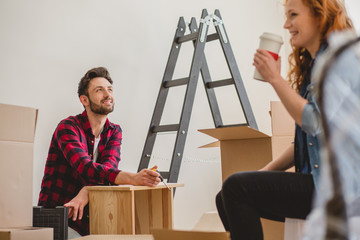 Smiling man assembling a cabinet and his girlfriend drinking coffee while furnishing interior