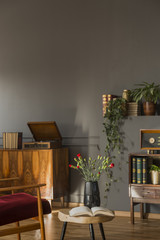 Flowers on table next to red armchair in grey flat interior with record player on cabinet. Real...