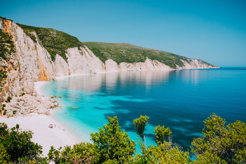 Sunny Fteri beach lagoon with rocky coastline, Kefalonia, Greece. Tourists under umbrella chill relax near clear blue emerald turquoise sea water