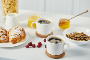 close up view of cups of coffee, croissants and raspberries for breakfast on white surface