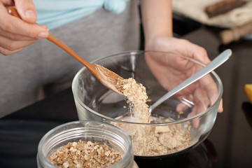 Close-up shot of crop hand using spoon to pour dry oatmeal into glass bowl while preparing breakfast in kitchen