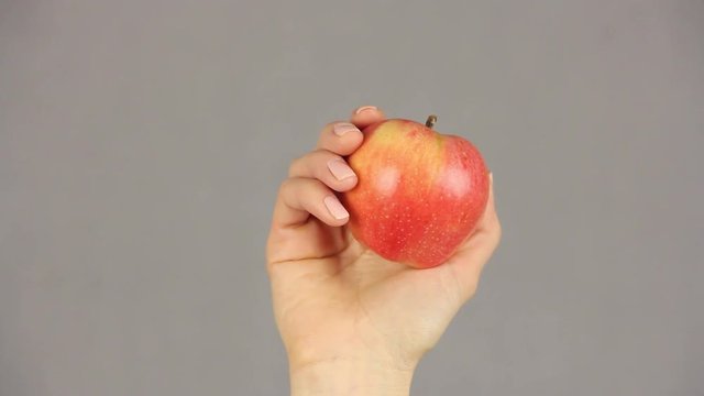 female hand holds an red apple on a gray background, rises from the bottom of the frame and drop down