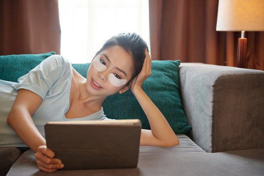 Pretty Asian female with under-eye patches smiling and browsing tablet while lying on comfortable couch in living room - Powered by Adobe