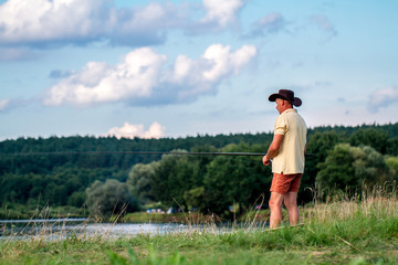 A fisherman in shorts, a hat and a T-shirt is fishing on the shore of the lake. Fishing, hobbies, recreation