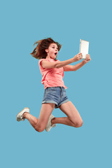 Jump of young woman over blue studio background using laptop or tablet gadget while jumping. Runnin...