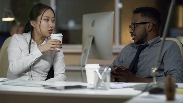 Medium shot of Asian woman and black man talking and drinking coffee when sitting at desk in office
