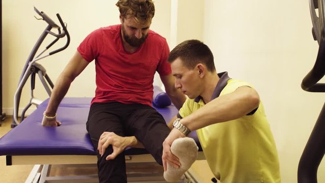 Doctor physiotherapist doing stretching exercises for young disabled man at the rehabilitation center. The patient's face expressing effort.