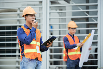 Side view of young Asian man in waistcoat and hardhat holding modern tablet and talking on radio while standing on construction site near colleague