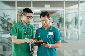 Two Asian health professionals in medical scrubs standing in hospital hall and browsing tablet together