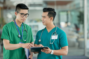 Two young Asian men in medical scrubs pointing at information on tablet screen while standing on...