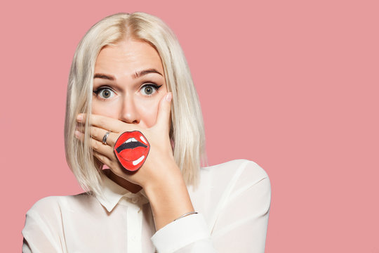 Young woman with the painted mouth on her hand. Studio portrait of blonde hair model on pink background. Emotion of surprise and shock. Concept of fun and tomfoolery.