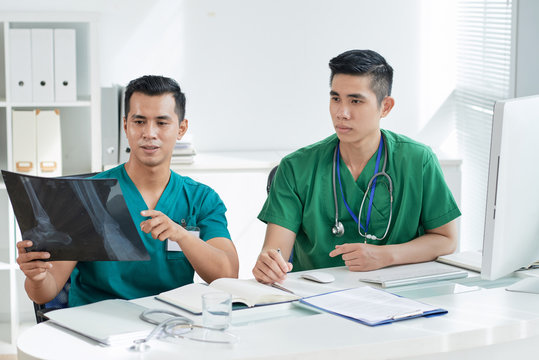 Two Asian men in scrubs looking at bone X-ray picture while sitting at desk in hospital office
