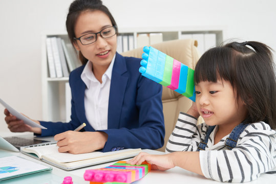 Charming Asian girl with colorful blocks sitting at working table with working woman in suit in office