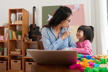 Asian woman with laptop working at home and asking playful little daughter to be quiet 