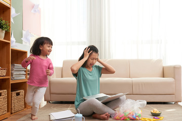 Stressful Asian woman with laptop sitting on floor and pressing head with noisy girl running around 