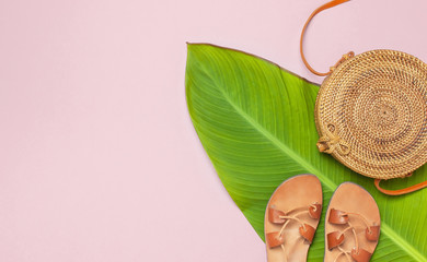 Fashionable natural round rattan bag, summer leather sandals and tropical leaves on pink background flat lay. Top view with copy space. Trendy bamboo bag Ecobags from Bali. Summer fashion concept.