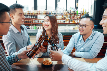 Asian cheerful people toasting with beer bottles while smiling and communicating in leisure spending time in bar 