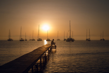 Obraz premium Man silhouette sitting on the wooden pier on the sea bay with sailing boats on the horizon. Breathtaking golden sunset.