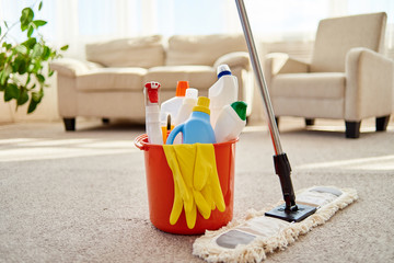 Cleaning set for different surfaces in orange bucket and mop on floor in living room, copy space. Cleaning service concept. Bucket with cleaning items for kitchen, bathroom or toilet