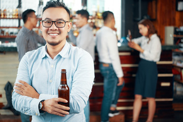 Smiling Asian man in glasses and shirt holding beer bottle and looking at camera in bar with people on background
