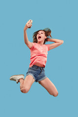 always on mobile. Full length of pretty young woman taking phone while jumping against blue studio...