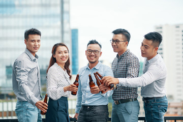 Group of Asian men and woman standing together and clinking with beer bottles smiling at camera on...