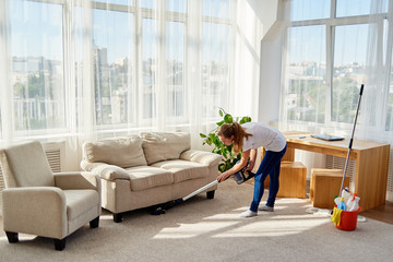Full body portrait of young woman in white shirt and jeans cleaning carpet with vacuum cleaner in...