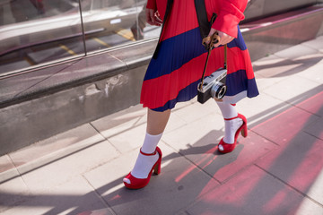 Elegant lady wearing stylish red skirt and shoes holds in hands vintage photo camera. Low angle...