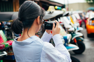 Asian woman traveler uses digital camera takes photo with colorful Tuk tuks car background- Bangkok-Thailand- Asia