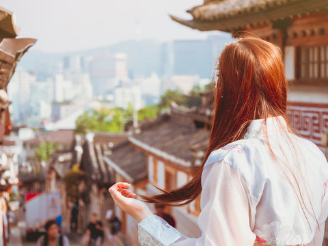 travel and tourist asia concept from backside of beauty long hair woman in korea traditional cloth (hanbok) stand, take picture and see to people in vintage town with soft focus background