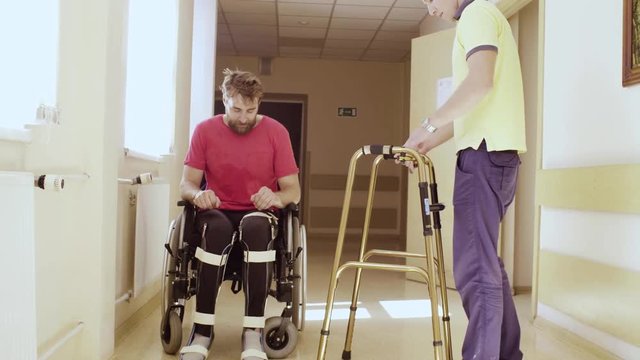 A tired young disabled man sitting in a wheelchair after walking on a walker. The doctor folds the walkers and takes him away.