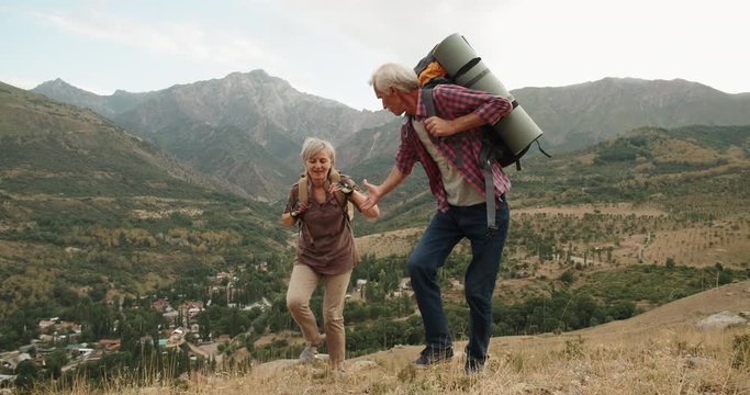 Mature caucasian senior on a hiking adventure taking wife's hand helping her climb up a mountain. tourism concept 4k