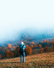 Naklejka premium Photographer taking photo of autumn landscape with foggy peaks and orange trees. Ukrainian Carpathians mountains