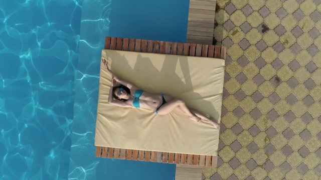 aerial, attractive girl in blue swimsuit and spectacles lies near a swimming pool at summer day