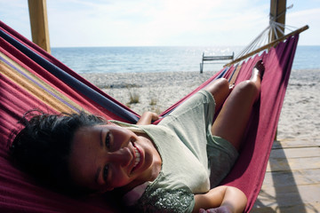 brunette girl lies in a hammock and smiling on the beach with sea as background