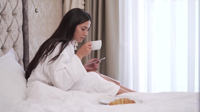 A young brunette is sitting on bed looking at the phone, drinking coffee or tea. She puts the cup back and types something.