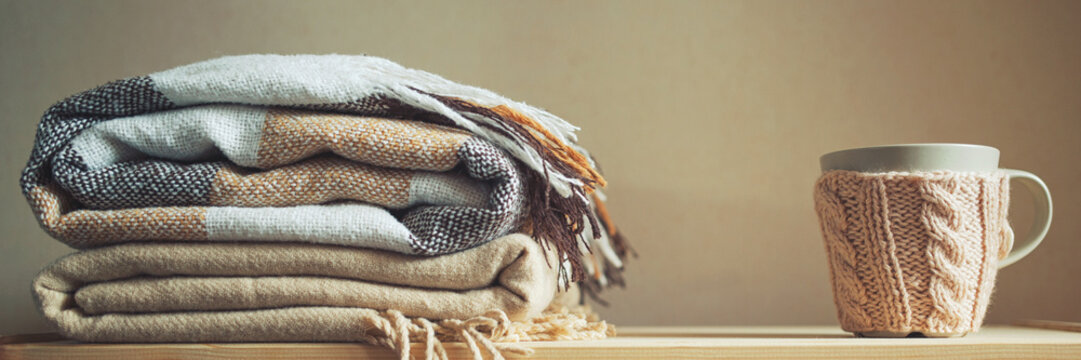 stack of beige checkered wool blankets and cup on a wooden chest. autumn - winter concept of home warmth and comfort. toned, long banner